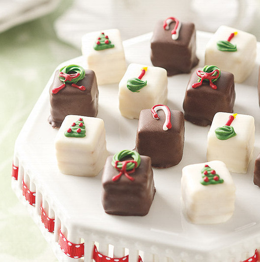 White and milk chocolate petit fours decorated with Christmas trees, wreaths, candles and candy canes on a white platter