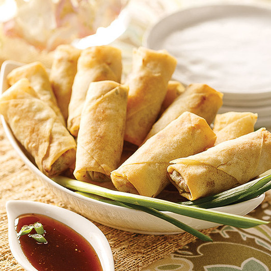 Golden vegetable spring rolls on a white platter beside a small dish of brown dipping sauce and green onions