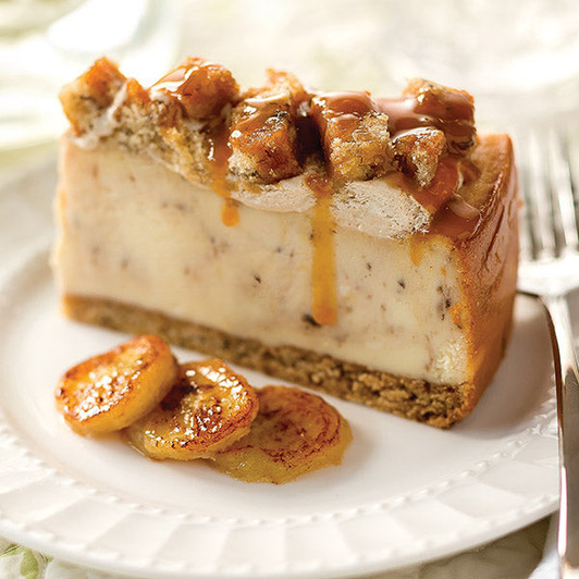 Bananas Foster Cheesecake slice with banana cake base, creamy filling and caramel-drizzled topping; fruit garnish is a serving suggestion.