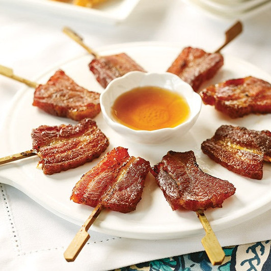Skewered bacon pieces arranged around a bowl of glaze on a white plate.