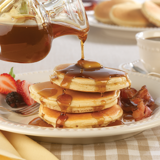 Maple syrup poured over pancakes, with bacon as a serving suggestion