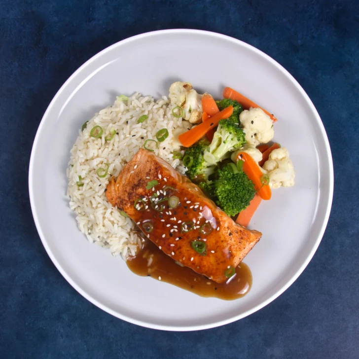 Prepared Asian sesame salmon meal with sesame seeds, white rice, broccoli, cauliflower, and carrots.