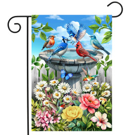 Vibrant garden flag with colorful birds, flowers, and butterfly near birdbath under blue sky with clouds.