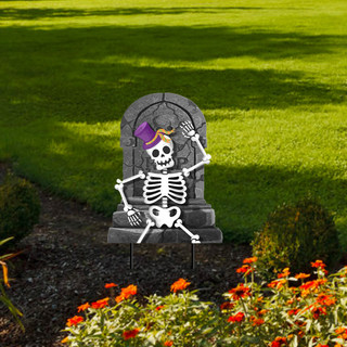 Metal skeleton wearing purple hat on gray tombstone in garden with orange flowers and green grass.