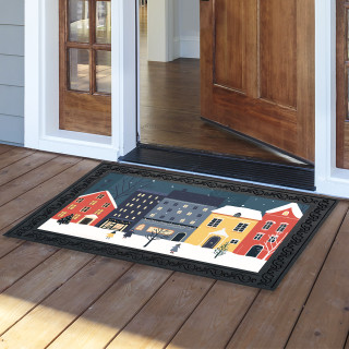 Snow Day Doormat with colorful winter village scene and black border on wooden porch outside doorway.