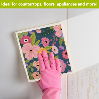 Hand in pink glove wiping white surface with Happy Blooms Swedish Dishcloth featuring floral design.