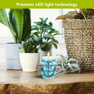 Welcome Anchor blue flameless candle with nautical design on wooden table surrounded by plants.