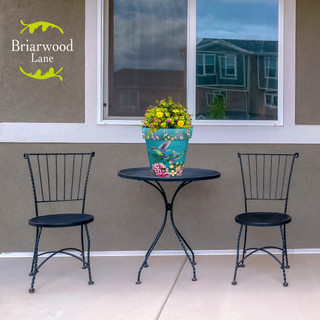 Springtime Hummingbirds Medium Planter on a small black metal table between two matching chairs outdoors.