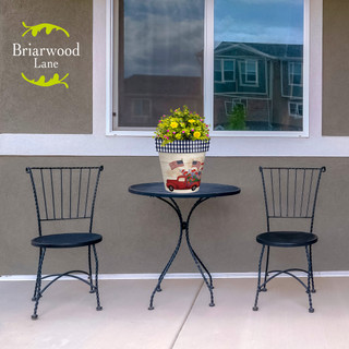 Stars and Stripes Truck Medium Planter with yellow flowers on outdoor table between two black chairs.