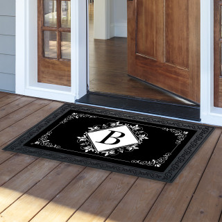 Black and white monogram doormat with decorative border and letter B on wooden porch floor.