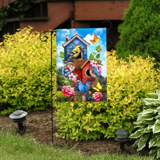 Vibrant garden flag with floral birdhouses and colorful birds on a decorative stand in a garden.