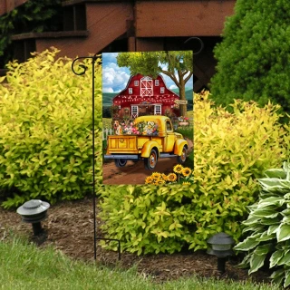 Garden flag with yellow truck and red barn surrounded by flowers and greenery outdoors.