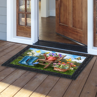 In the Garden Doormat with colorful floral design and black border on wooden porch outside doorway.