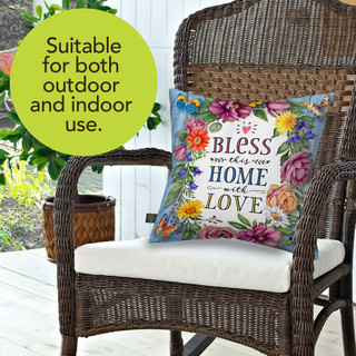 Bless This Home With Love Indoor/Outdoor Pillow on wicker chair with floral design and white cushion.