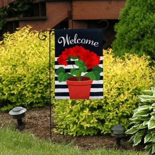 Red geraniums in red pot on black and white garden flag with welcome text on burlap.
