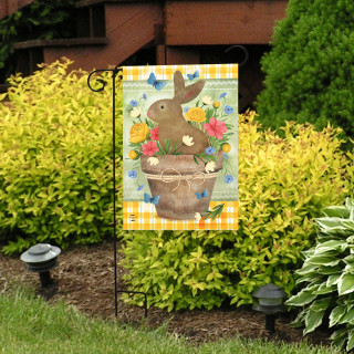 Brown bunny in a flowerpot with colorful flowers on a yellow and white garden flag outdoors.
