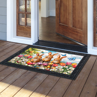 Bunny Greeting Doormat with colorful floral design and two brown bunnies on wooden porch outside doorway.
