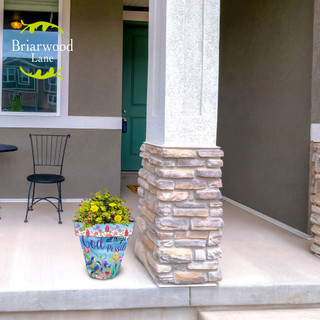 Multicolored With God Medium Planter with yellow flowers on a house porch next to a pillar.