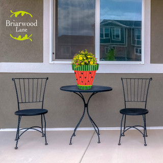 Briarwood Lane Watermelon Medium Planter with yellow flowers on outdoor table between two black chairs.