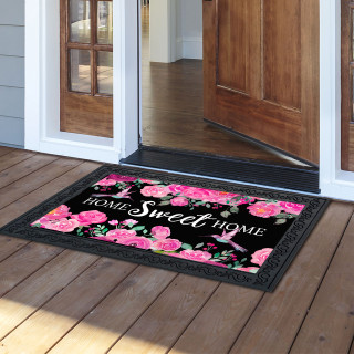 Black doormat with pink hummingbirds, roses, and "Home Sweet Home" text on wooden porch floor.