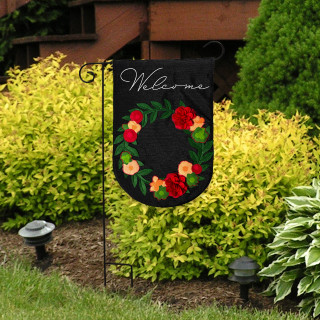 Black garden flag with colorful floral wreath and "Welcome" text on burlap material outdoors.