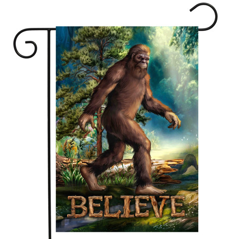 Believe Bigfoot garden flag with brown Sasquatch walking in forest landscape with trees and water.