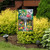 Briarwood Lane Birdbath in Bloom garden flag with vibrant flowers and birds on a decorative stand outdoors.