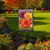 Checkered garden flag with vibrant orange, yellow, and purple mums and welcome text on stand outdoors.