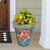 Large teal planter with colorful flower design holding green plants with yellow and pink flowers.