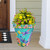 Large turquoise planter with dragonfly design, colorful flowers, and greenery, on a wooden porch floor.