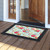 Spring Floral Doormat with black border on wooden porch outside open doorway with pink flowers.
