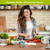 Woman preparing salad in kitchen with Summer Gnomes flameless candle on table nearby.