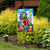 Vibrant garden flag with floral birdhouses and colorful birds on a decorative stand in a garden.