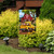 Garden flag with yellow truck and red barn surrounded by flowers and greenery outdoors.