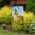 Garden flag with cross, sunflowers, and "God Bless Our Home" text on a garden stand outdoors.