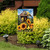 Garden flag with goldfinches, sunflowers, and birdhouse on a decorative stand in a garden setting.