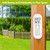 White Home Metal Thermometer on a wooden post with green wreath and black lettering outdoors.