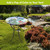 Country Birds Glass Birdbath on a garden table surrounded by lush greenery and vibrant flowers outdoors.