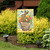 Brown bunny in a flowerpot with colorful flowers on a yellow and white garden flag outdoors.