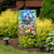 Vibrant garden flag with colorful birds, flowers, and birdbath design on a decorative stand outdoors.