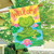 Peaceful Frog House Flag with green frog, pink flowers, and welcome text on colorful background.