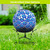 Blue and white scalloped mosaic gazing ball with solar LED lights on a black metal stand outdoors.