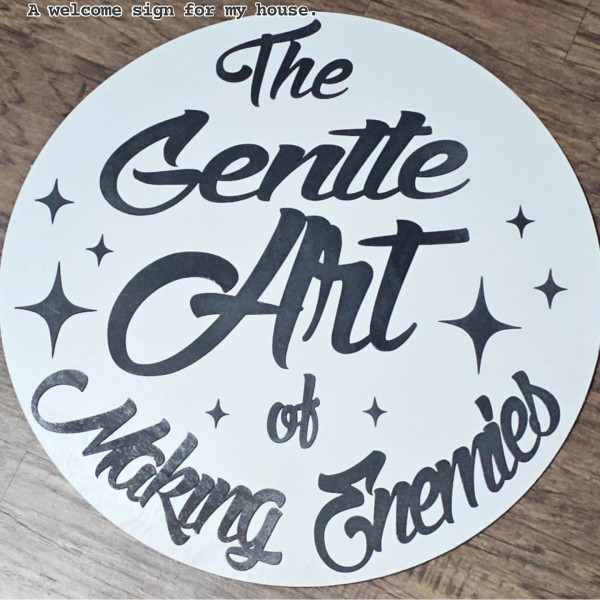 Round 18-inch wooden sign with the quote “It’s the Gentle Art of Making Enemies” in black cutout letters on a matte white background, modern and bold wall decor.