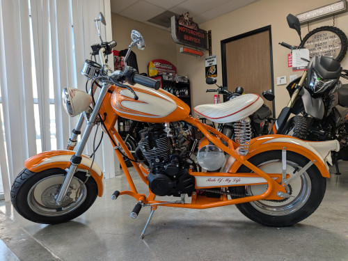Left-side view of the "Creamsicle" CSC motorcycle in a showroom, highlighting the "Ride Of My Life" swingarm graphic and chromed components.