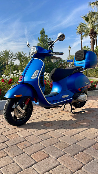 A Matte Blue 2020 Vespa GTS 300 HPE parked on a brick walkway, showing the LED headlight, chrome mirrors, red front suspension spring, and the integrated rear storage box with a passenger backrest.
