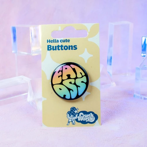 1.25 inch round button with bold rainbow text reading 'EAT ASS' on a black background, queer pride accessory by Crushworthy.