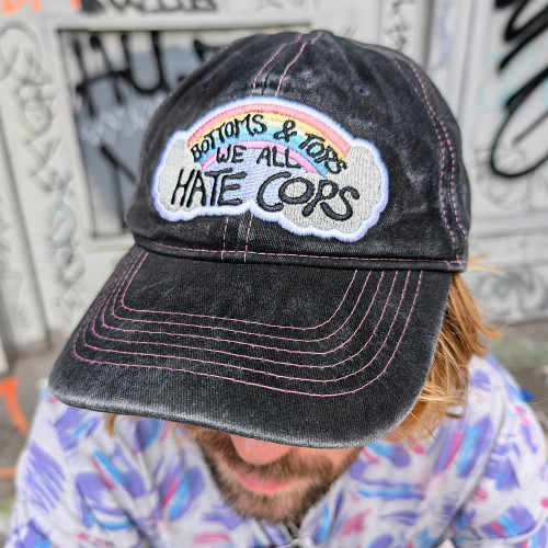Bottoms & Tops We All Hate Cops Cap by Crushworthy - Pastel embroidery that says "Bottoms & Tops We All Hate Cops" on top of a rainbow with clouds below it, embroidered directly onto an acid wash black baseball cap.
