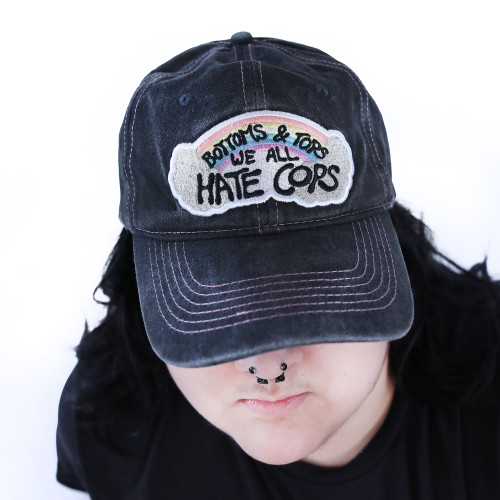 Bottoms & Tops We All Hate Cops Cap by Crushworthy - Pastel embroidery that says "Bottoms & Tops We All Hate Cops" on top of a rainbow with clouds below it, embroidered directly onto an acid wash black baseball cap.
