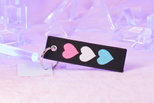 Thriving Despite It All keychain by Crushworthy - Trans pride colored hearts on one side, and text reading "thriving despite it all" on the other of a durable embroidered keychain