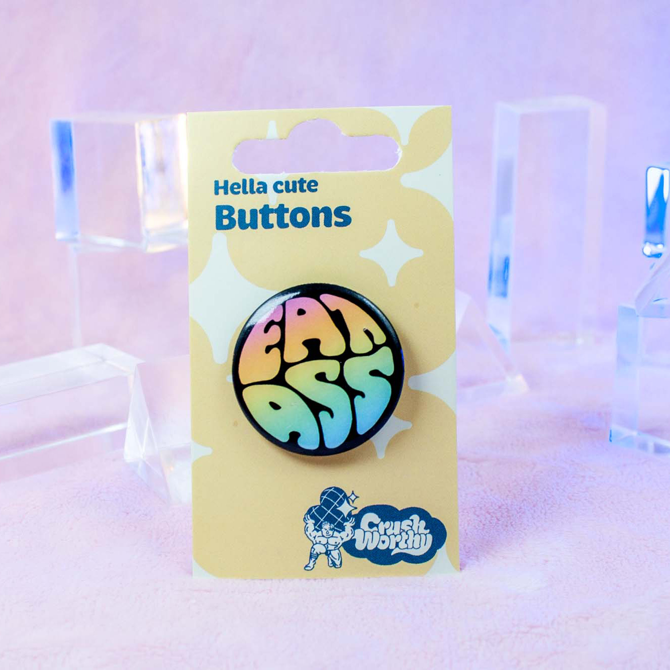 1.25 inch round button with bold rainbow text reading 'EAT ASS' on a black background, queer pride accessory by Crushworthy.